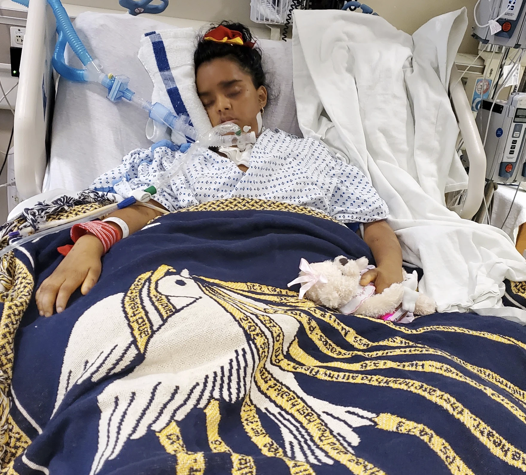 Danielle in the ICU holding her teddy bear, beside her Isaiah 40:31 angel-embroidered blanket.